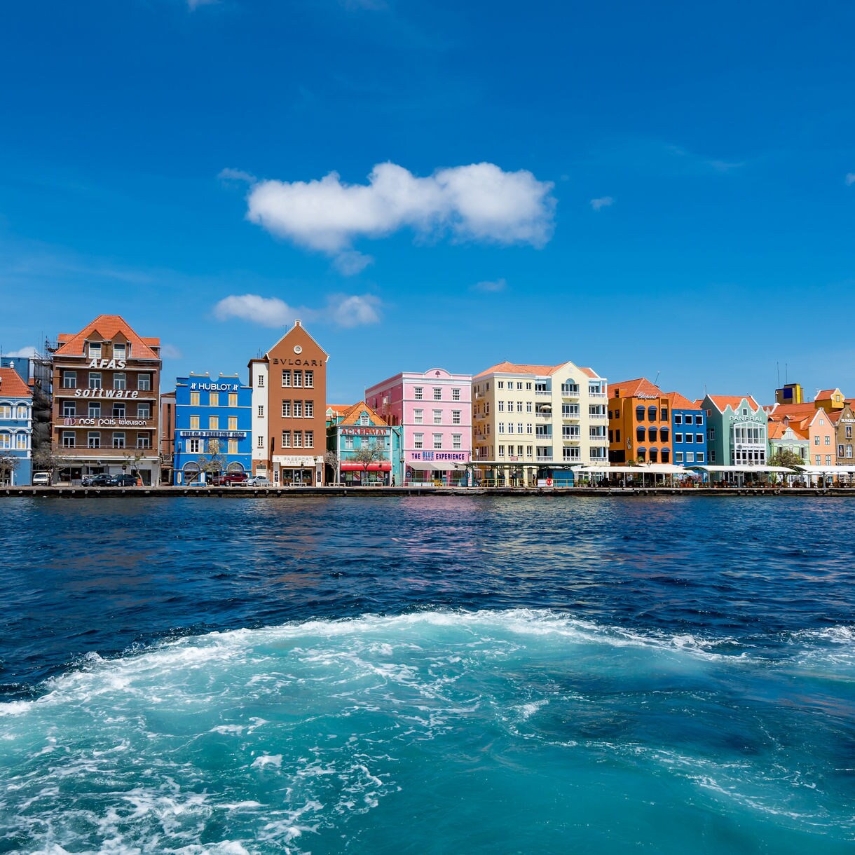 iew from a boat's wake of a colorful row of Dutch colonial buildings lining the waterfront of Willemstad Curaçao under a blue sky with scattered clouds.