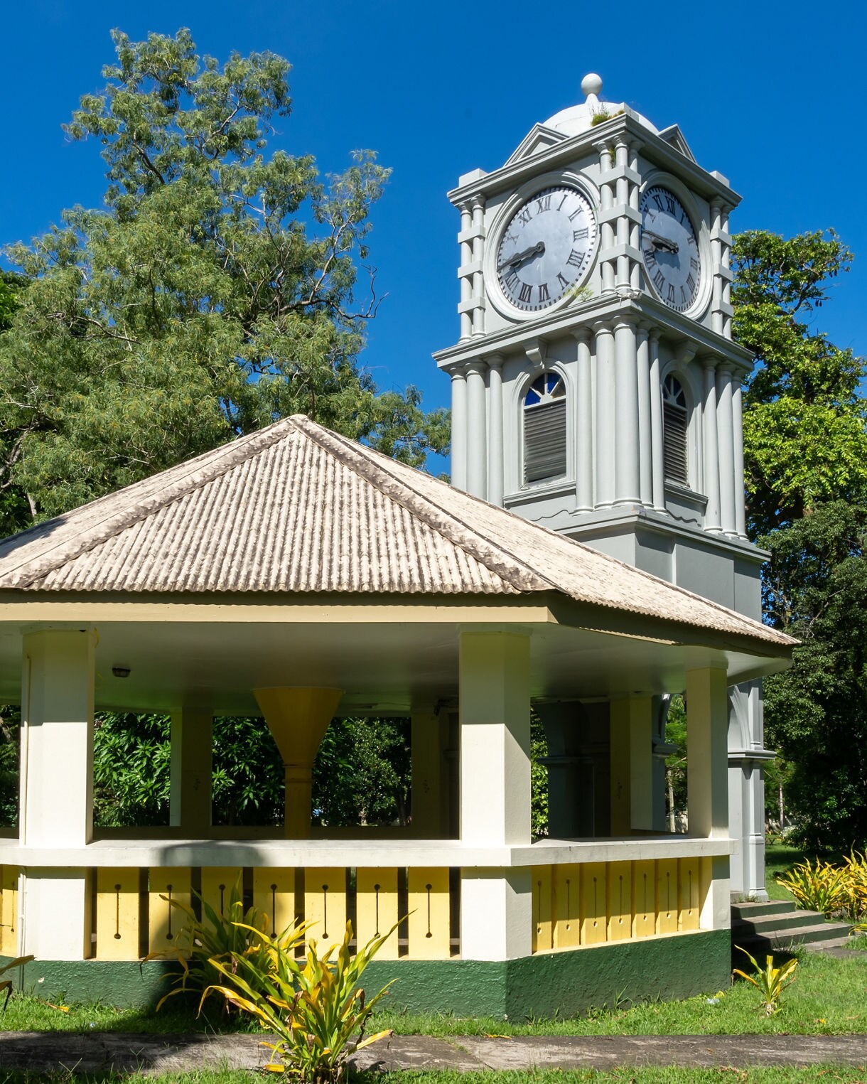 A pale gray historic clock tower rising behind a small open-air pavilion surrounded by vibrant green tropical foliage in Thurston Gardens, Suva.