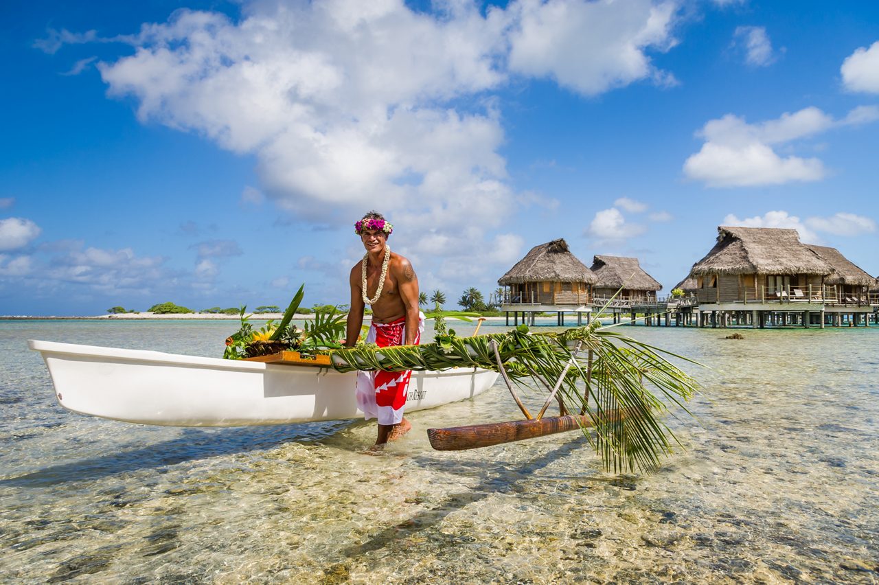 A Tahitian man dressed in traditional attire stands with an outrigger canoe filled with tropical greenery and food, on the shallow, clear waters of Tikehau Island in Tahiti, with overwater bungalows in the background.