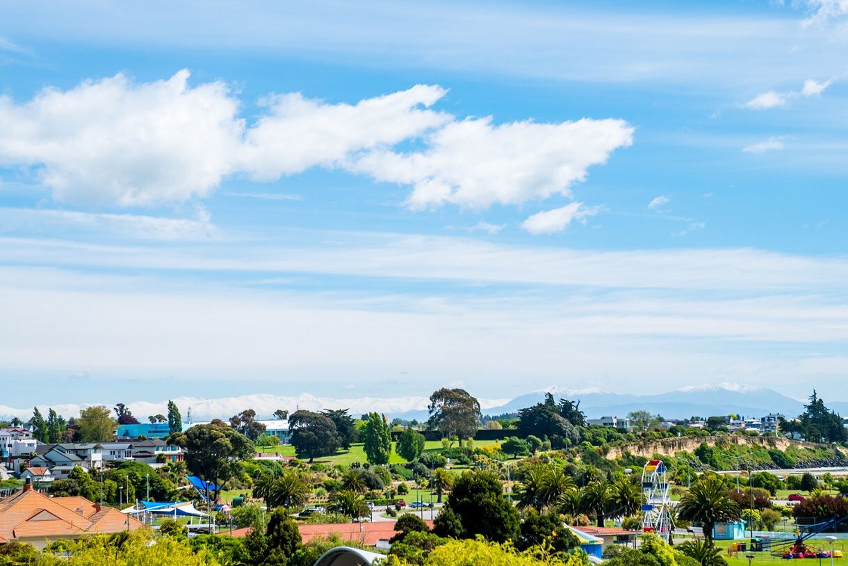 Bright view of Timaru, New Zealand showing leafy neighborhoods, parkland and distant snow-covered mountains under a clear blue sky.
