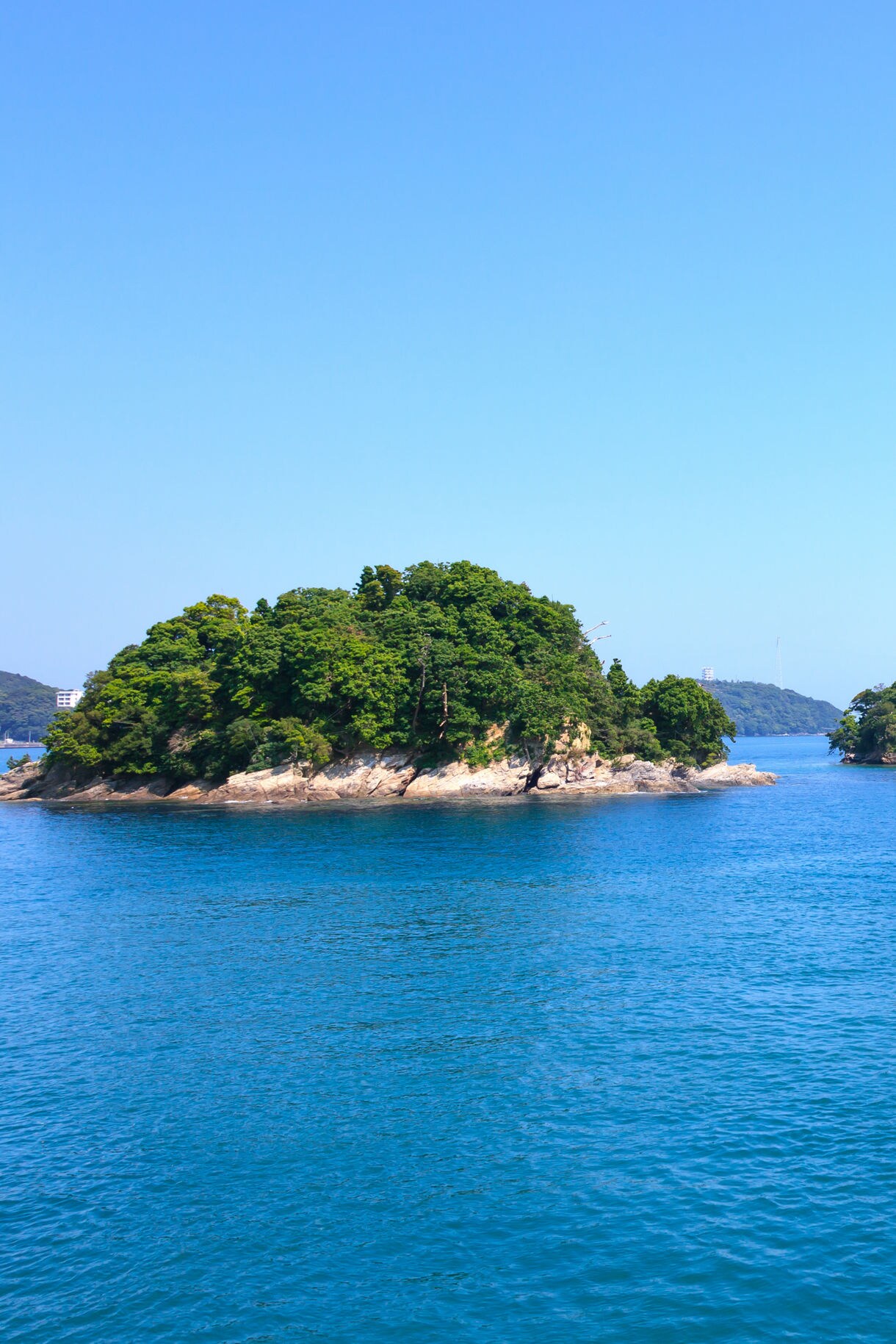 View through a bright blue architectural frame showing a small tree-covered island surrounded by deep blue water under a clear sky, with white building elements visible at the edges.