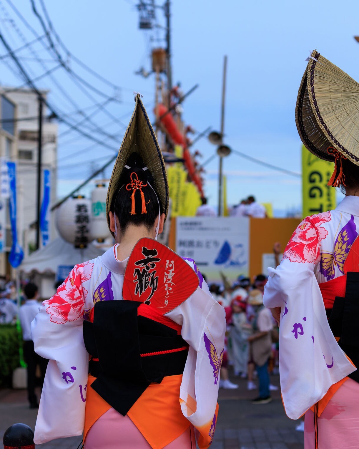 Back view of two women in yukata and straw hats at a Tokushima street festival with crowds and lanterns.
