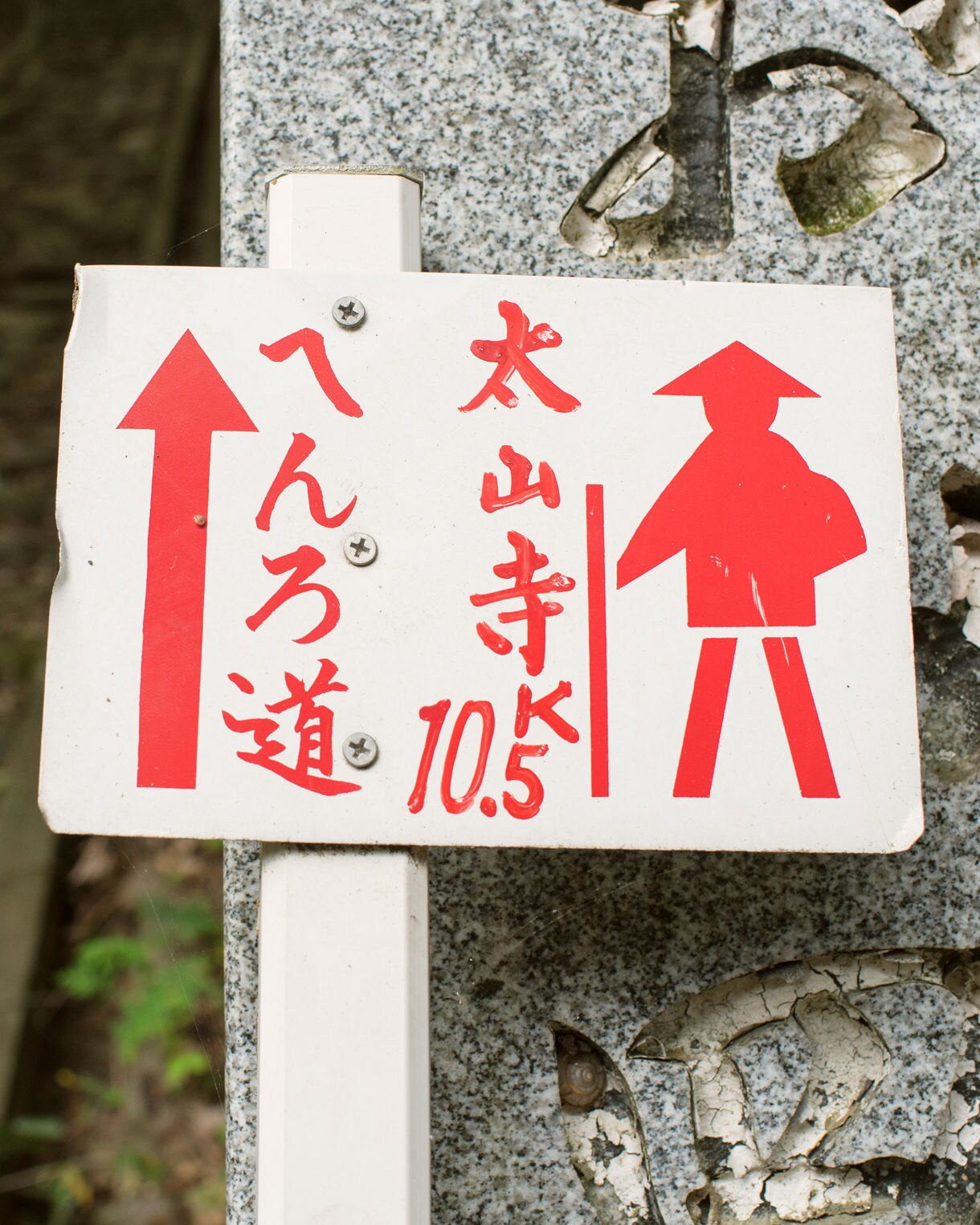 Close-up of a Japanese pilgrimage sign with red text and arrows, set beside stone steps in a forest.