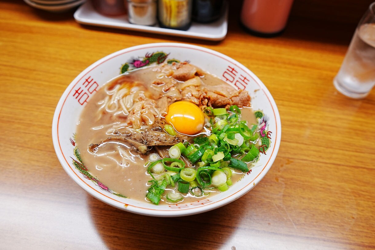Bowl of Tokushima ramen with pork slices, raw egg, green onions and rich broth on a wooden counter.