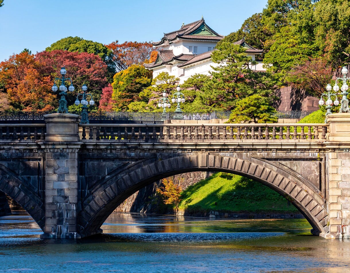 A stone bridge over a calm moat leading toward the Imperial Palace, surrounded by colorful trees, manicured greenery, and traditional palace buildings under a clear blue sky.