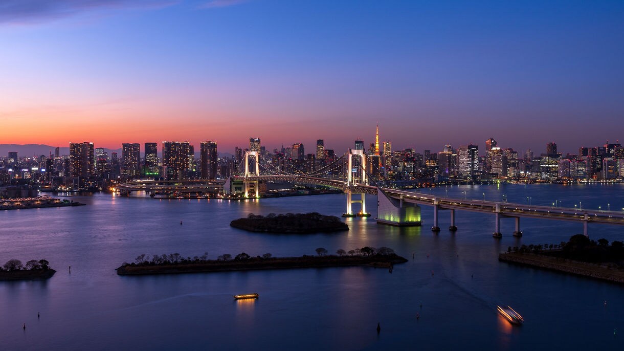 A breathtaking twilight panorama of Tokyo's illuminated skyline reflected across Tokyo Bay, with the iconic Rainbow Bridge and Tokyo Tower glowing against a gradient sunset sky.