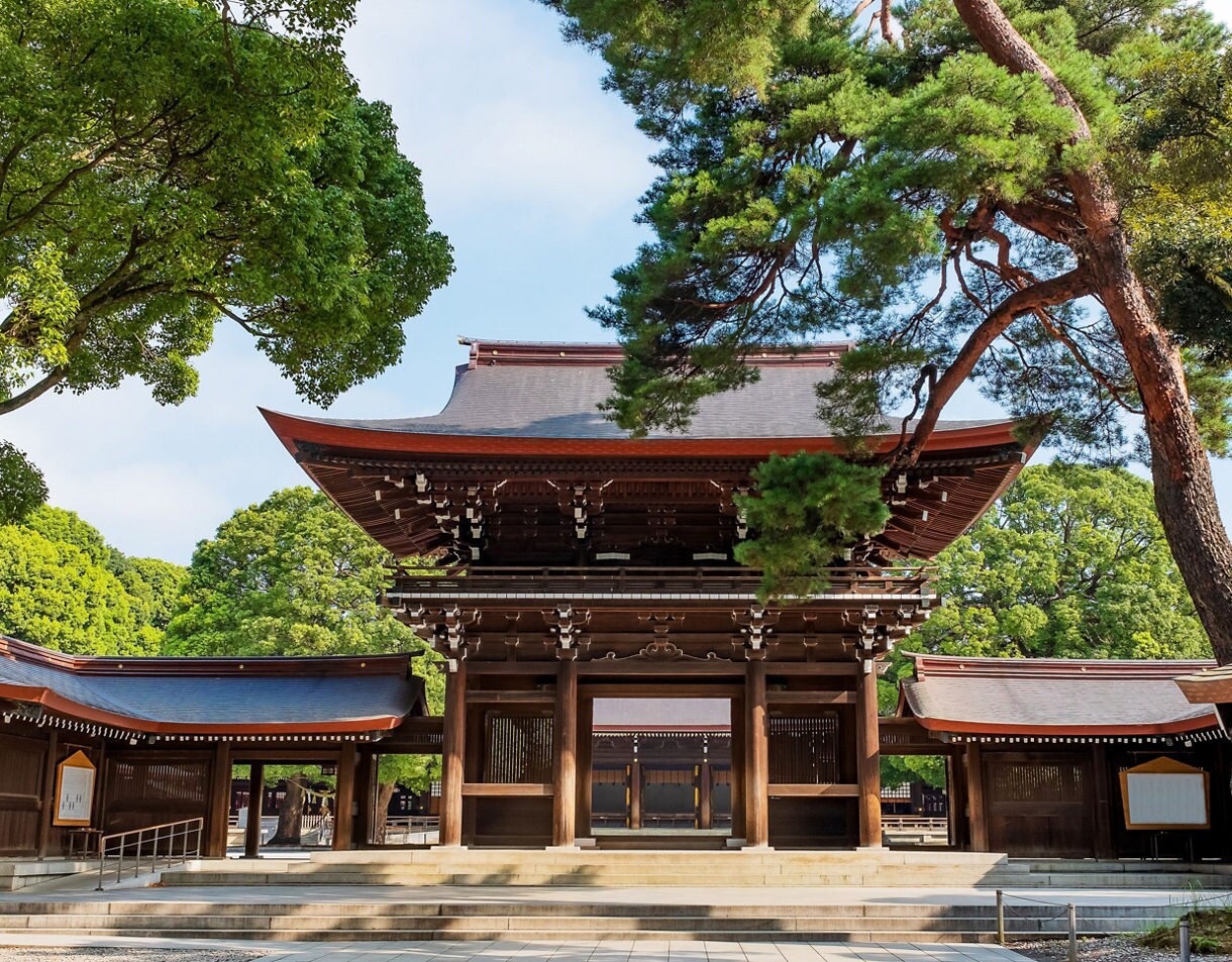 A large wooden entrance gate at Meiji Shrine surrounded by lush green trees and traditional structures under a bright blue sky.
