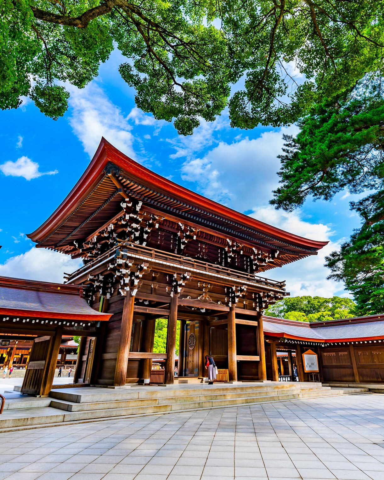 A large wooden entrance gate at Meiji Shrine in Tokyo, Japan, framed by green trees and a vivid blue sky, with a visitor standing at the threshold of the structure.
