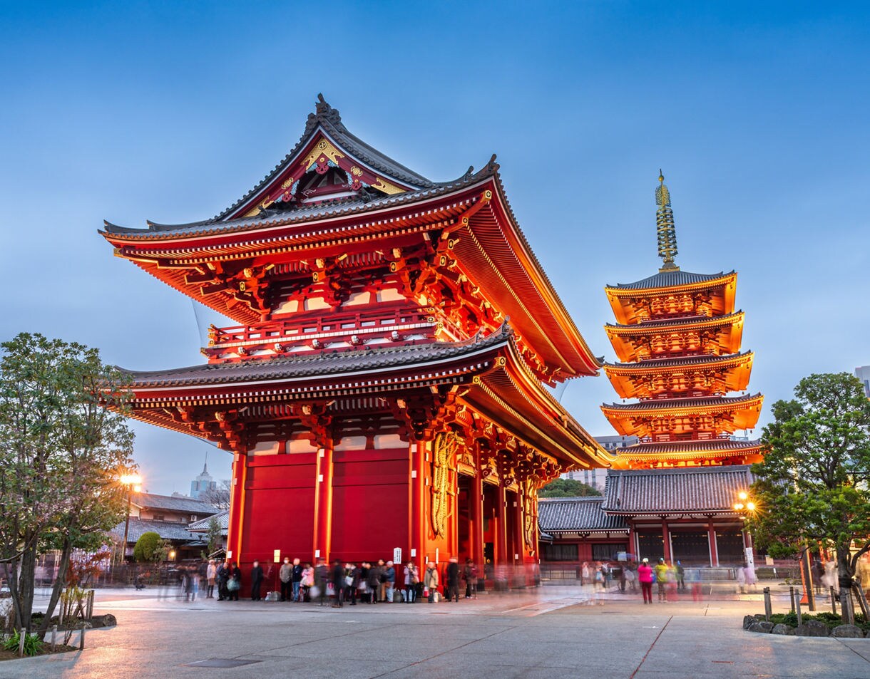 A bright red temple gate and illuminated pagoda at Sensoji Temple in Tokyo at dusk, with people walking and trees framing the scene.
