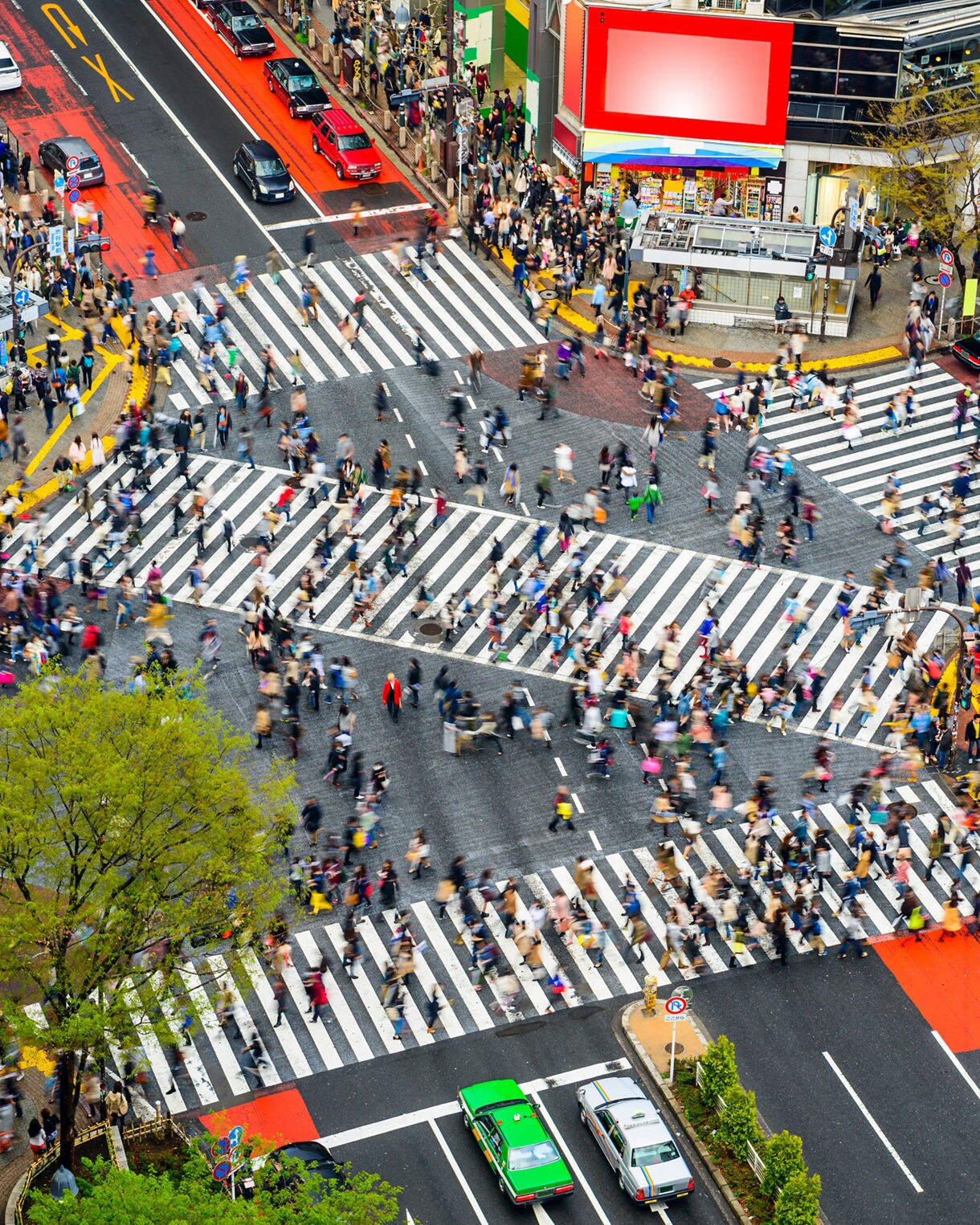 Aerial view of Shibuya Crossing in Tokyo, Japan, showing a large crowd of pedestrians moving in multiple directions across wide striped crosswalks surrounded by city buildings, taxis, and traffic.