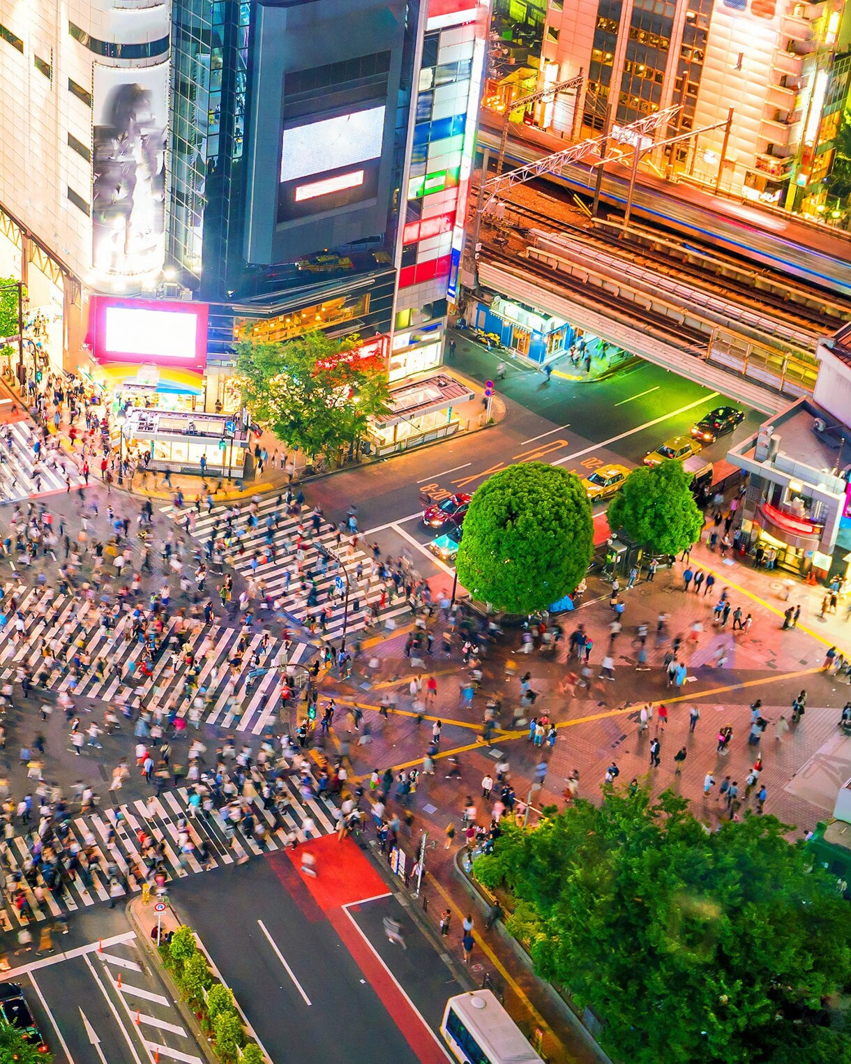 A high-angle view of Shibuya Crossing at night with colorful neon lights, crowded pedestrian crosswalks, and busy city streets filled with people and cars.