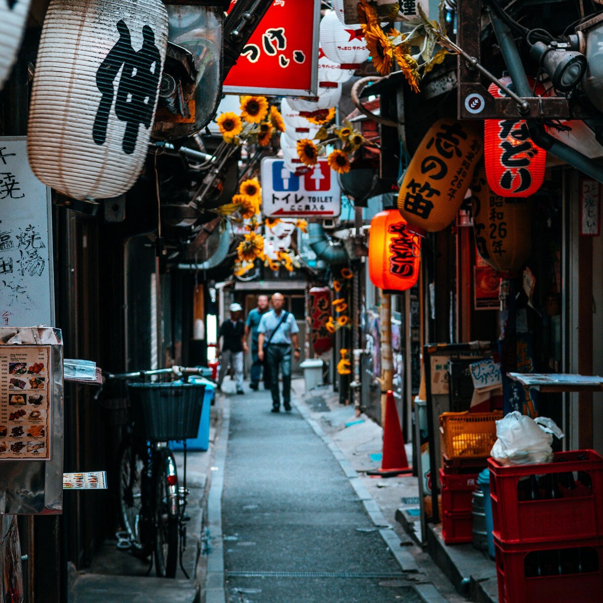 A narrow alleyway in Tokyo’s Golden Gai district, lined with small bars and eateries, glowing red and white lanterns, Japanese signage, and decorative sunflowers, with pedestrians walking down the path.