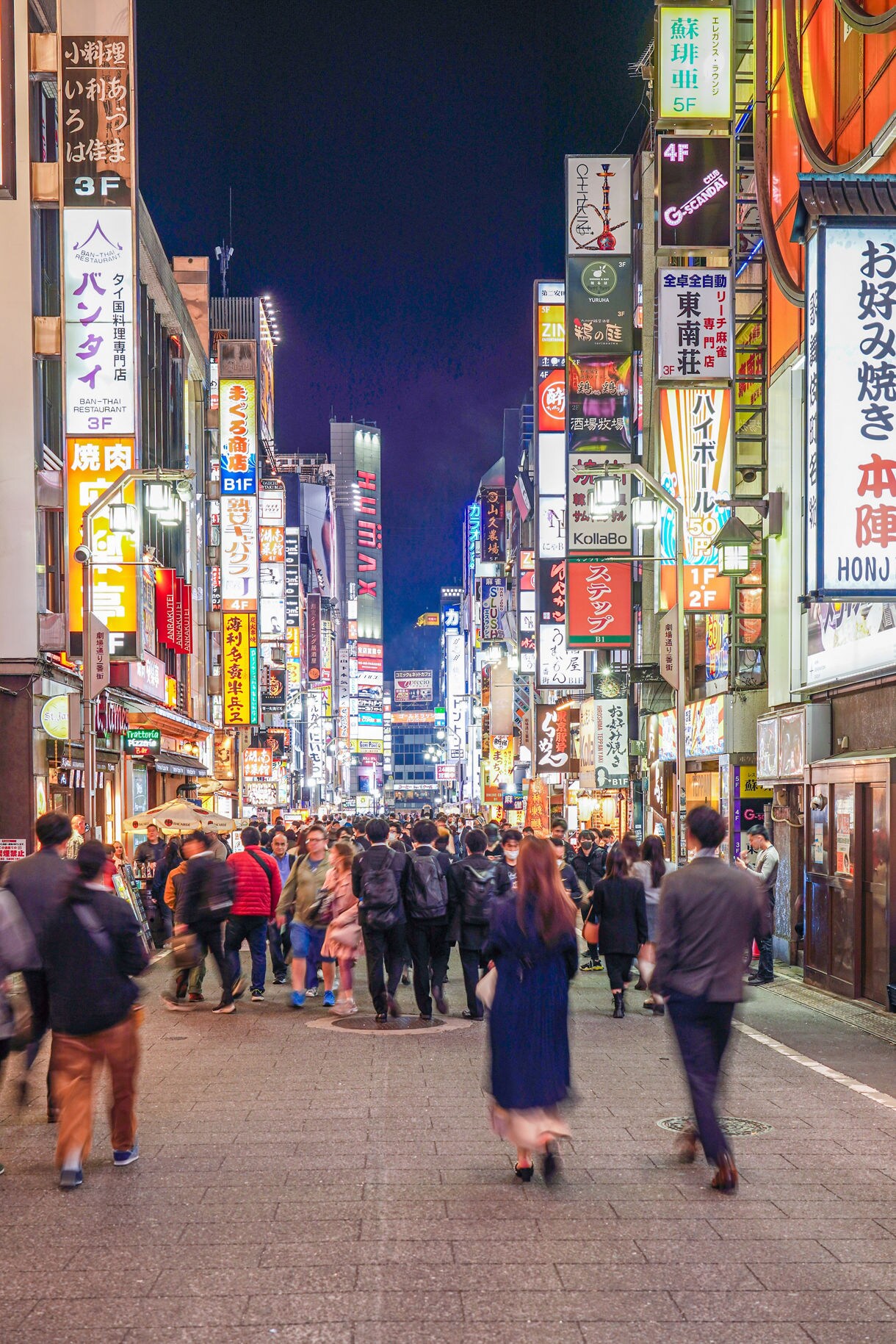 A busy Shinjuku street at night packed with people walking between tall buildings covered in bright neon signs and colorful advertisements.