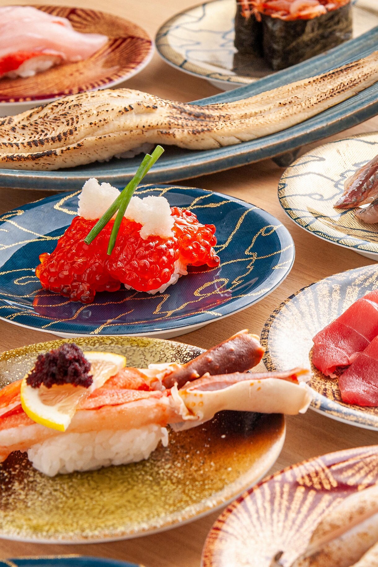Close-up view of various Japanese dishes on decorative plates, including bright orange salmon roe (ikura) on a blue plate, assorted sushi and sashimi pieces, with chopsticks and traditional serving dishes arranged on a table.