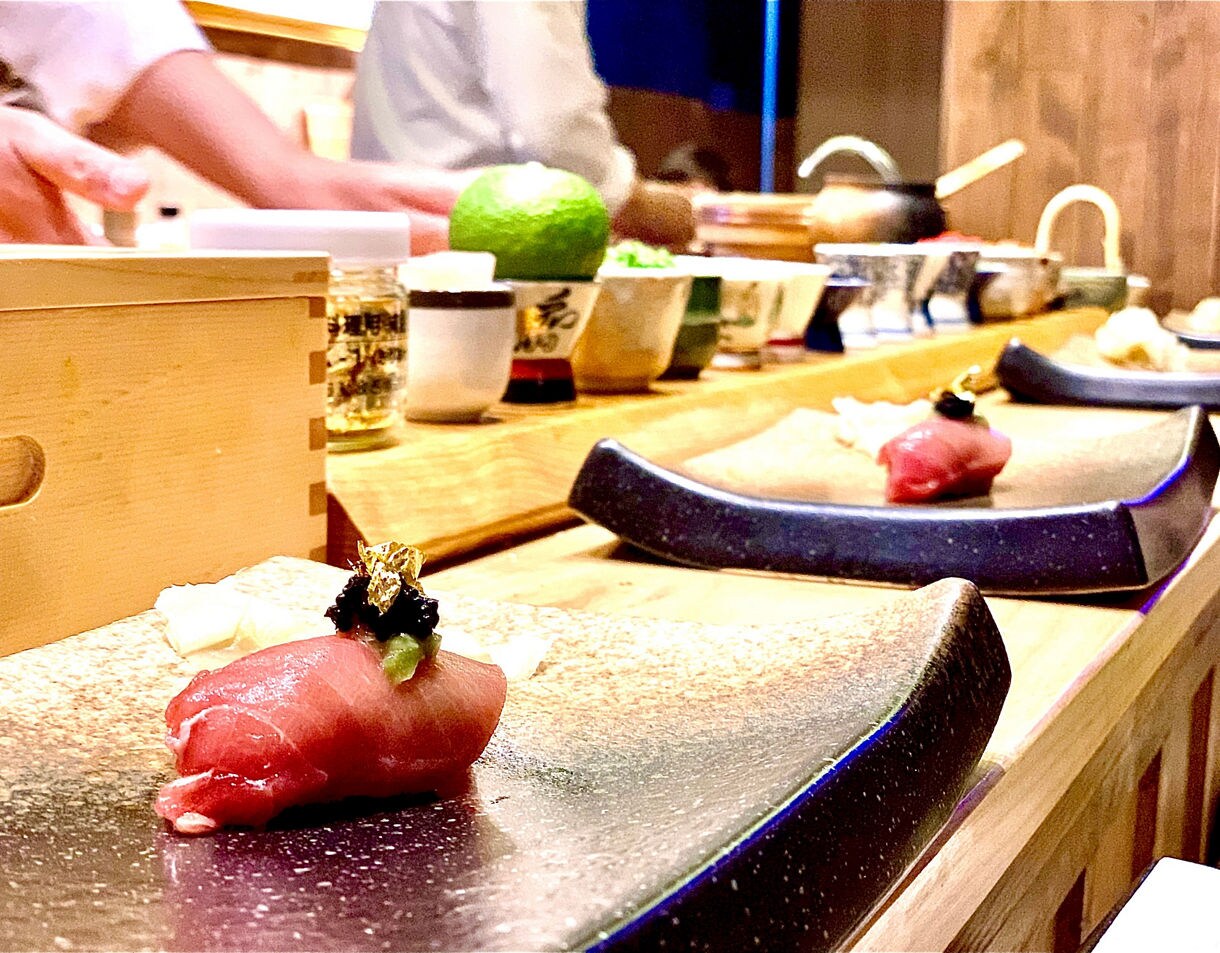 Close-up of a piece of sushi topped with garnish and gold flakes on a ceramic plate, with chefs preparing food and utensils arranged on a wooden counter in the background.