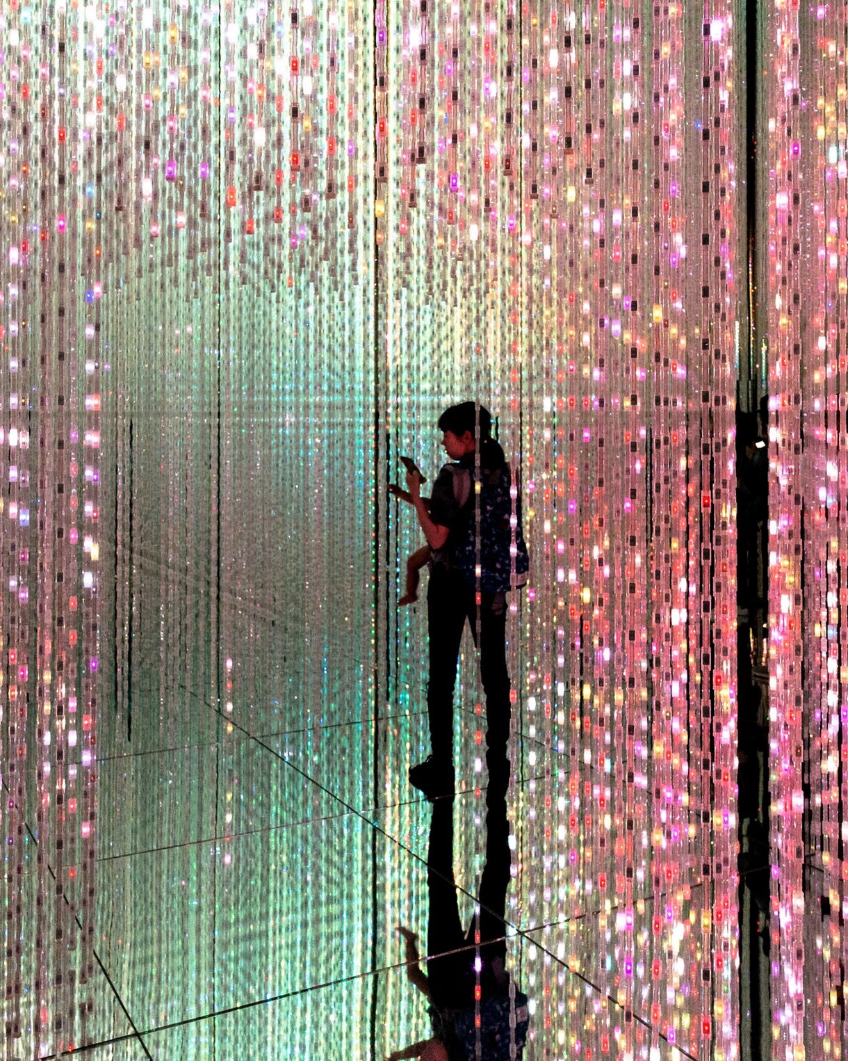 A person with a child walks through a mirrored room filled with hanging strands of tiny multicolored lights, creating glowing reflections and a tunnel-like effect.
