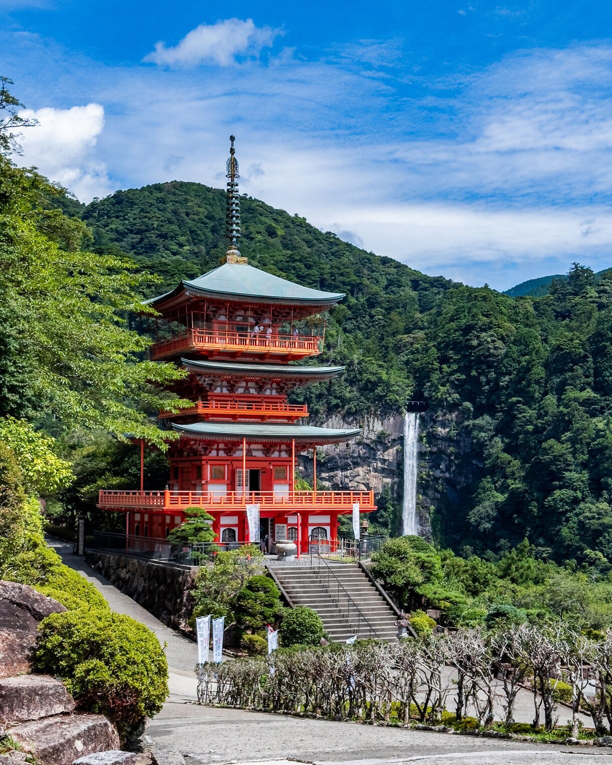 A red three-tier pagoda surrounded by lush green mountains with a tall waterfall cascading down a cliff in the background under a clear blue sky.