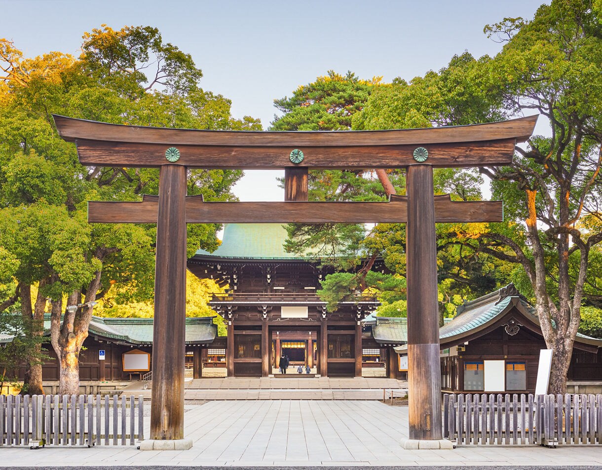 A large wooden torii gate leading to a traditional shrine courtyard, surrounded by tall green trees and historic wooden buildings under a clear sky.