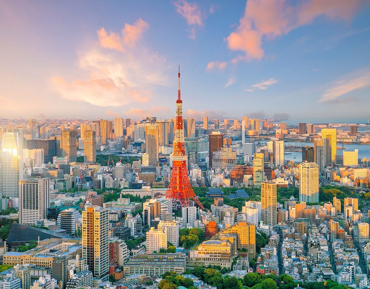 A sweeping panoramic view of Tokyo at sunset with Tokyo Tower in the center, tall buildings lit by warm sunlight, and the bay visible in the distance under a pastel sky.