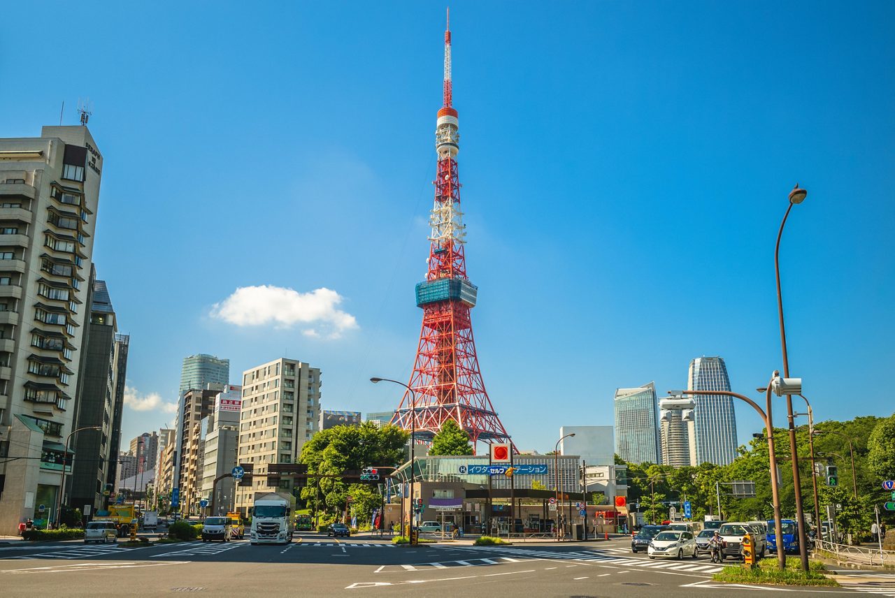 Daytime street-level view of Tokyo Tower, a red and white lattice structure surrounded by city buildings, traffic, and a bright blue sky with a few scattered clouds.