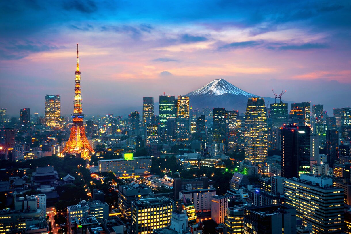 A view of Tokyo at twilight with Tokyo Tower glowing orange in the foreground, surrounded by illuminated skyscrapers and Mount Fuji snow-capped in the distance under a colorful sky.