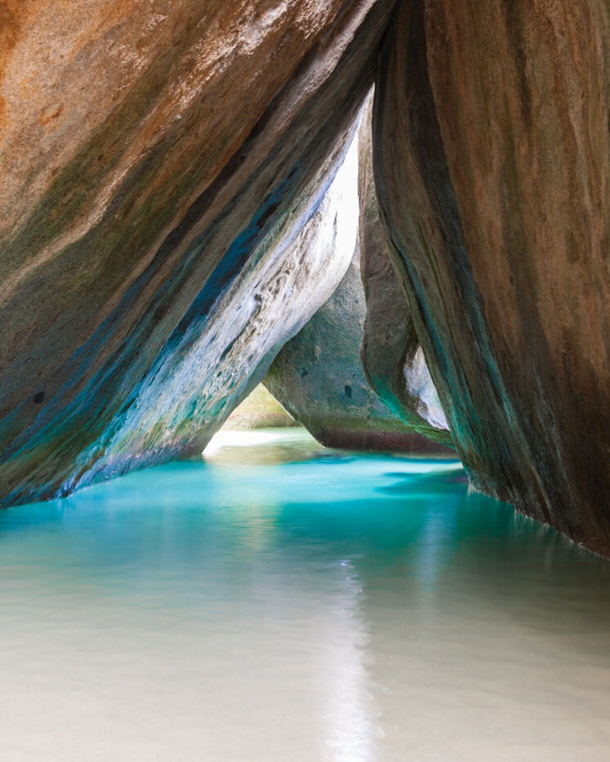 A narrow water passage at The Baths in Virgin Gorda featuring towering smooth granite boulders with streaks of green and orange, clear shallow water and sunlight streaming through gaps.