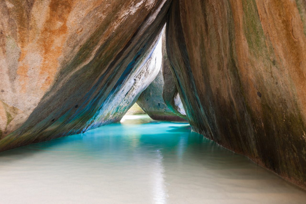 A narrow water passage at The Baths in Virgin Gorda featuring towering smooth granite boulders with streaks of green and orange, clear shallow water and sunlight streaming through gaps.
