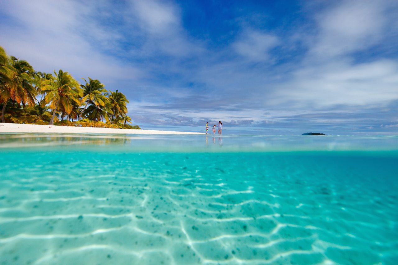 A serene view of Cane Garden Bay in Tortola with turquoise water, sandy beach and palm trees, while a family enjoys the calm, shallow water.