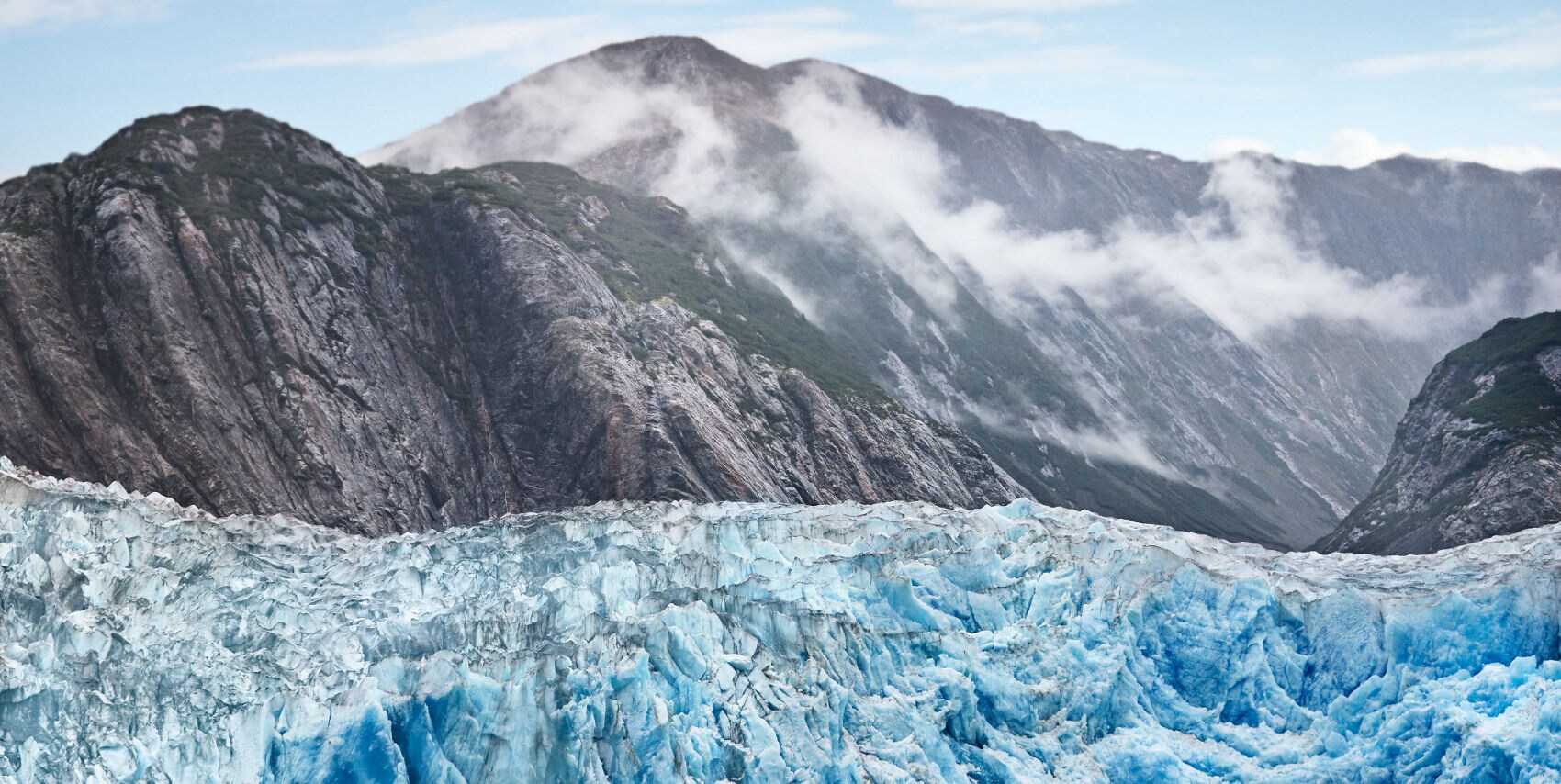 A massive blue glacier with crevassed ice face backed by steep dark mountain walls with mist drifting across the peaks in Endicott Arm, Alaska.
