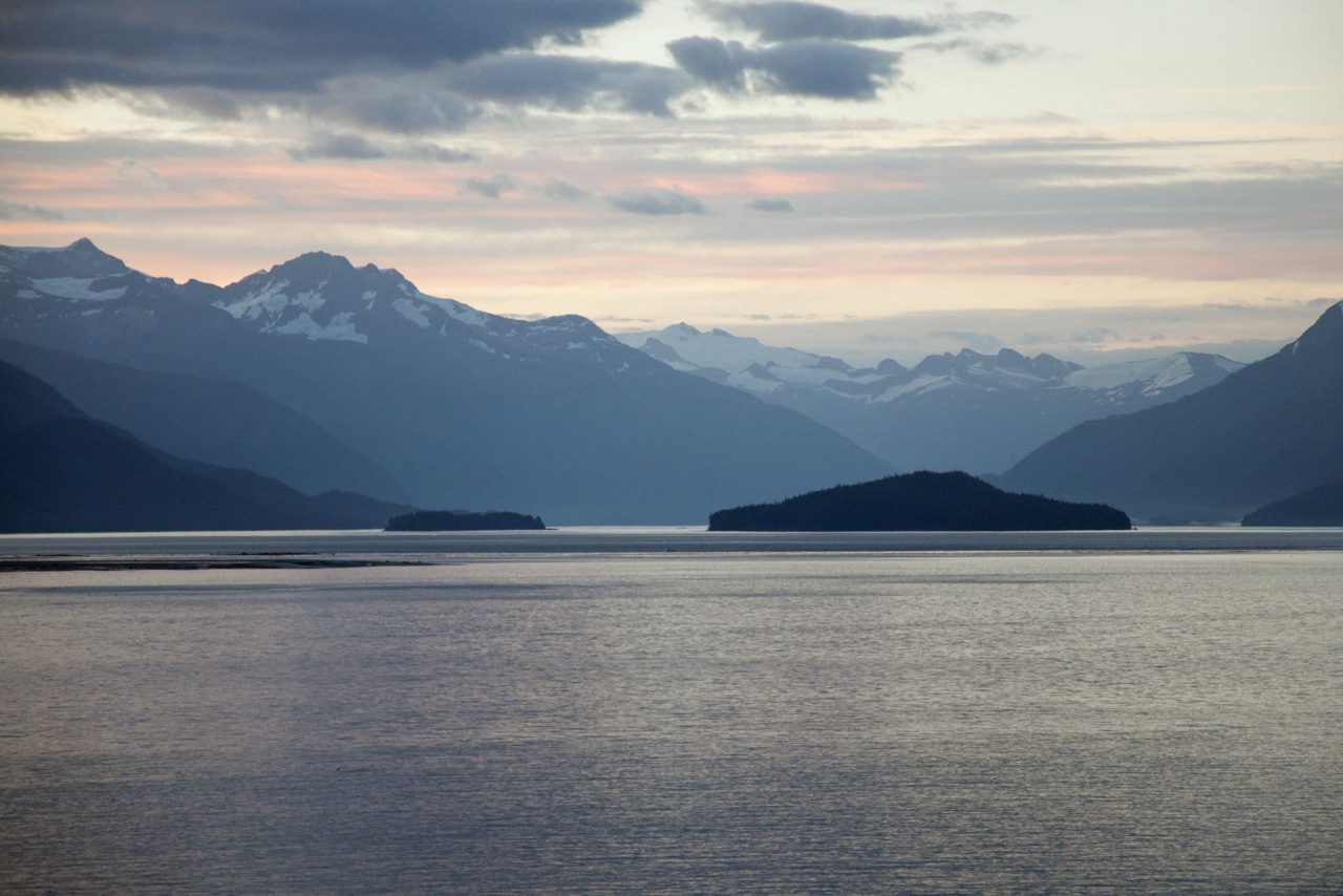 A serene fjord landscape with dark water, forested islands, and snow-capped mountains under a cloudy sky at dusk.