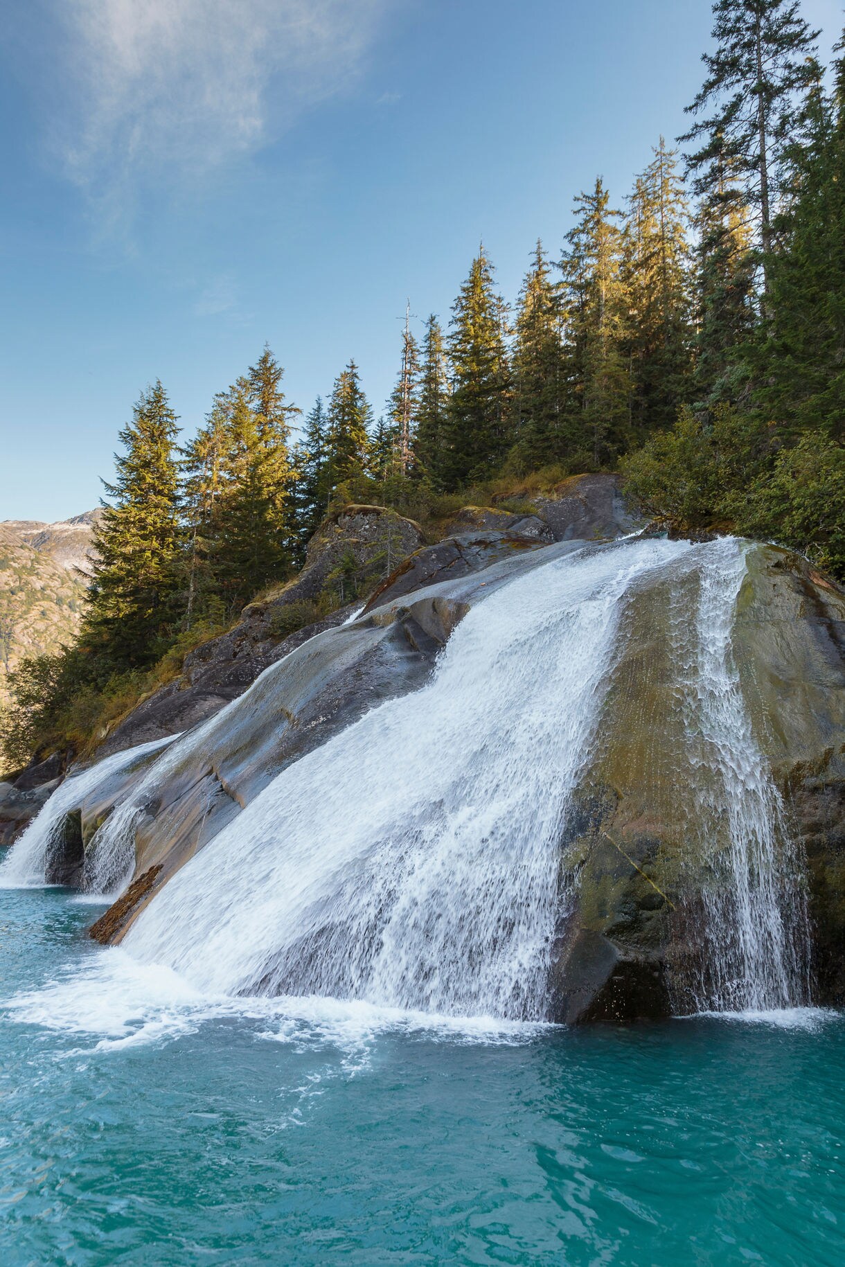 A cascading waterfall flowing over rounded rocks into bright turquoise water, surrounded by dense evergreen forest under a clear blue sky.