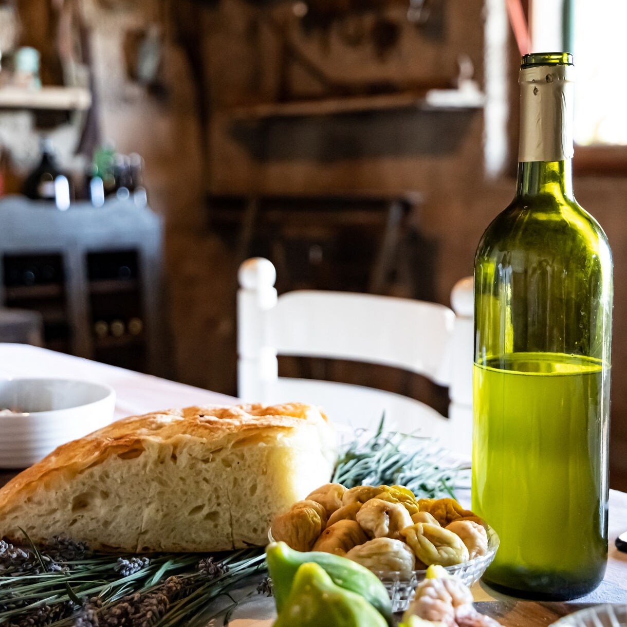 Hands-off table scene with rustic bread, cherry tomatoes, figs and a bottle of white wine in a cozy countryside dining room.