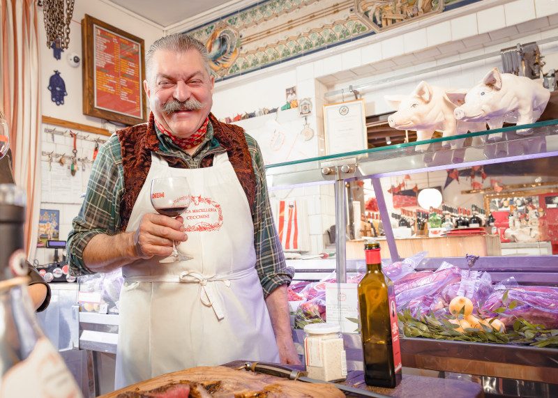 A butcher in traditional white apron smiling in his shop with tiled walls and hanging utensils.