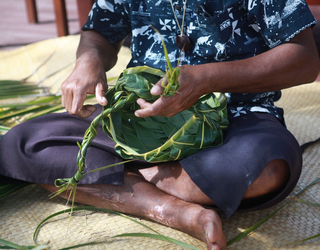 Close-up of a person weaving green palm leaves into a handmade basket while sitting on a woven mat.