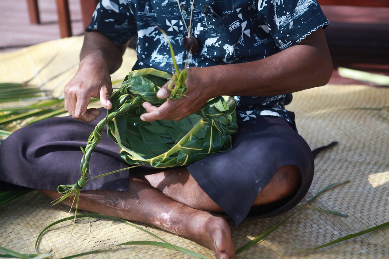 Close-up of a person sitting cross-legged on a mat weaving a traditional Fijian basket from green palm leaves.