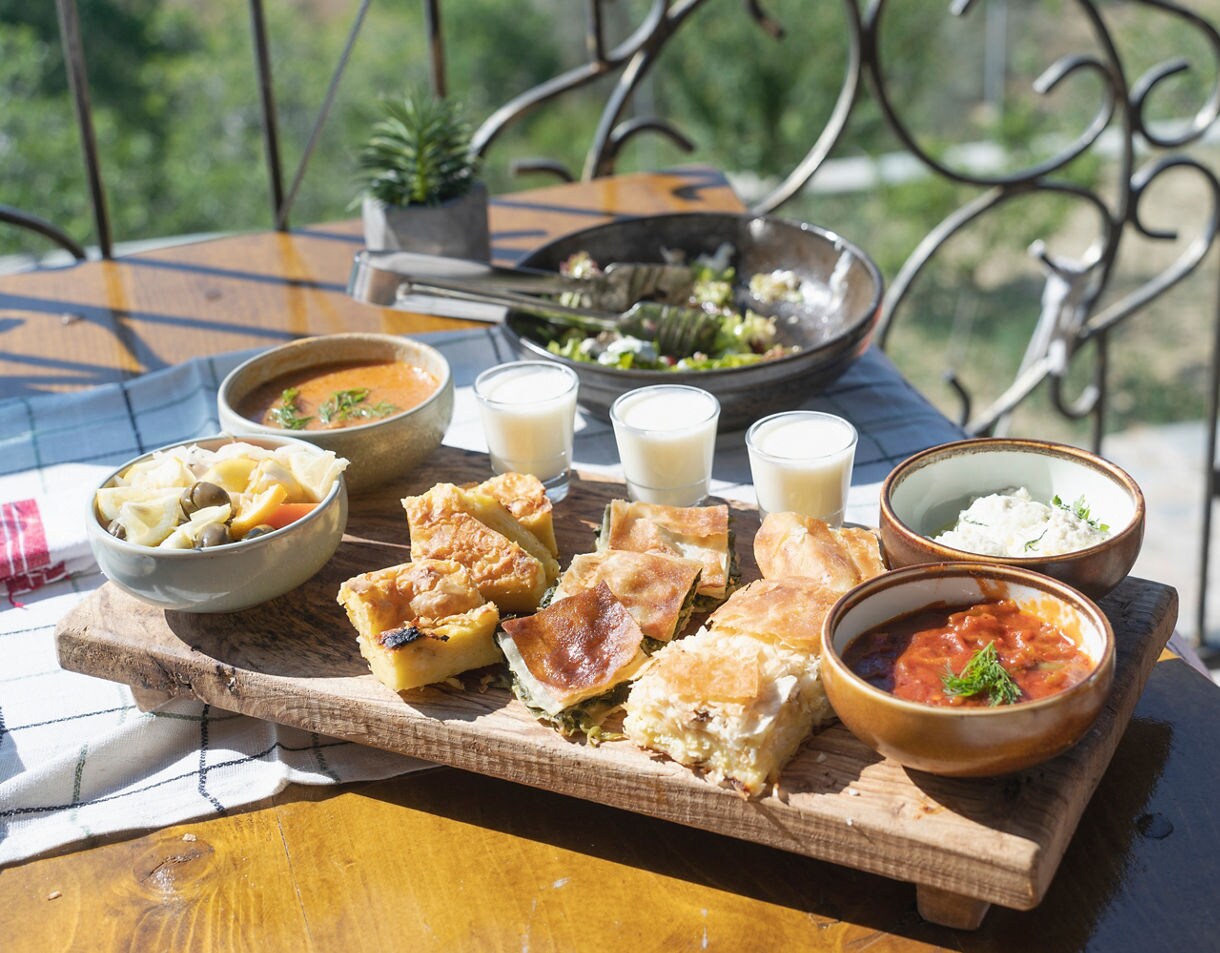 Wooden board filled with assorted Albanian pastries, vegetable stew, dips and small glasses of yogurt, surrounded by bowls of soup and salad on an outdoor table.