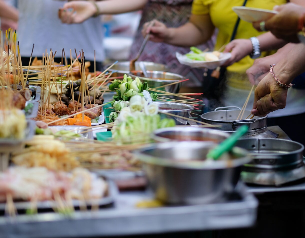 Close-up of lok lok skewers with vegetables and meats at a Penang street food stall, with hands dipping skewers into boiling pots.