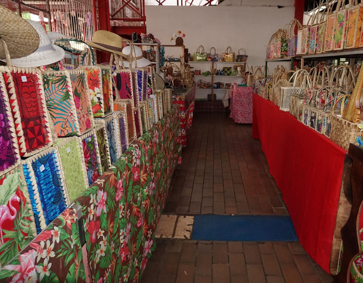 A narrow aisle inside a traditional Tahitian market lined with colorful woven bags, floral fabrics, straw hats and handmade crafts displayed on both sides.