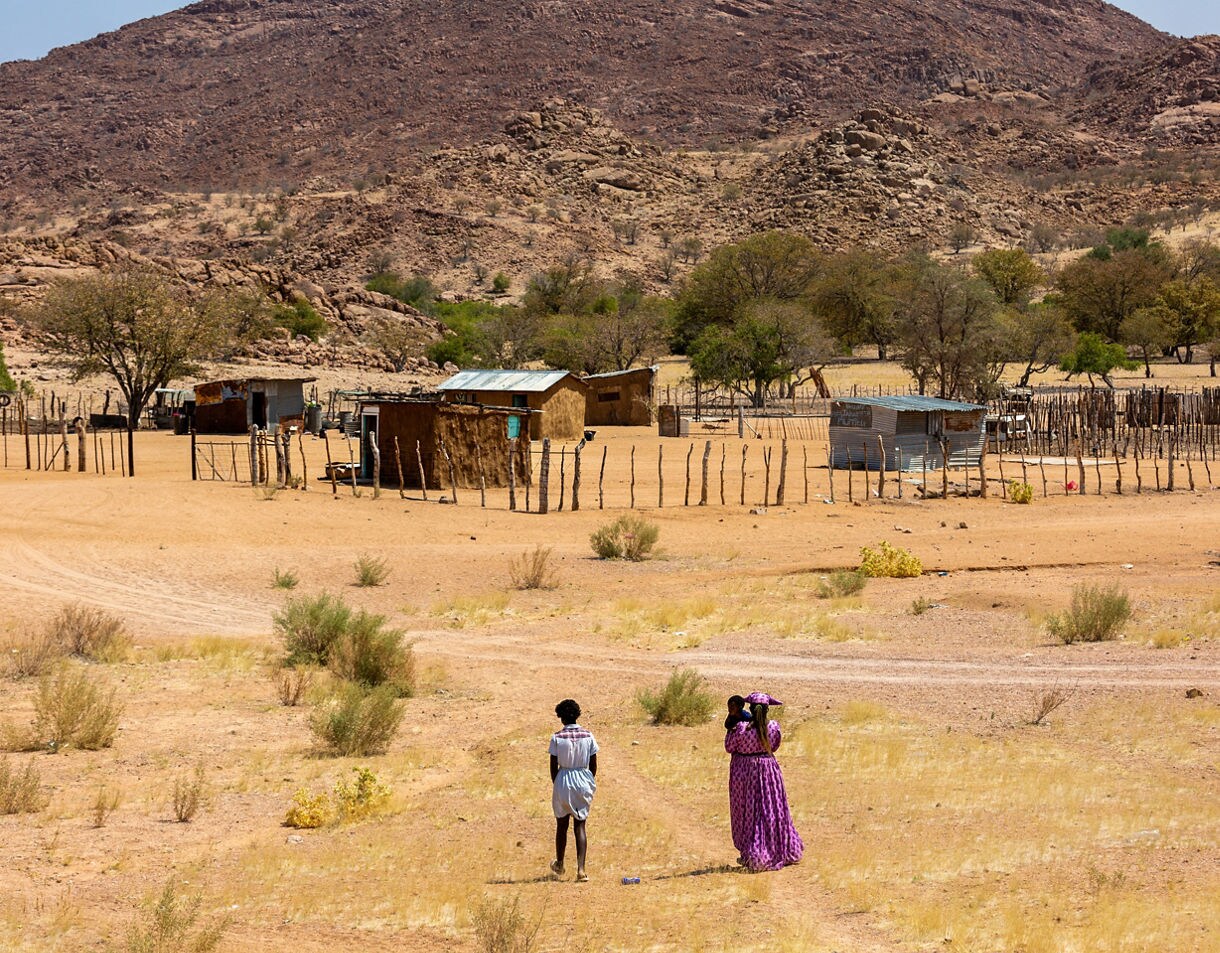 A woman in a bright purple dress and a child walk with another person toward a small rural village in Namibia, featuring simple wooden and metal homes surrounded by dry grassland and rocky hills.