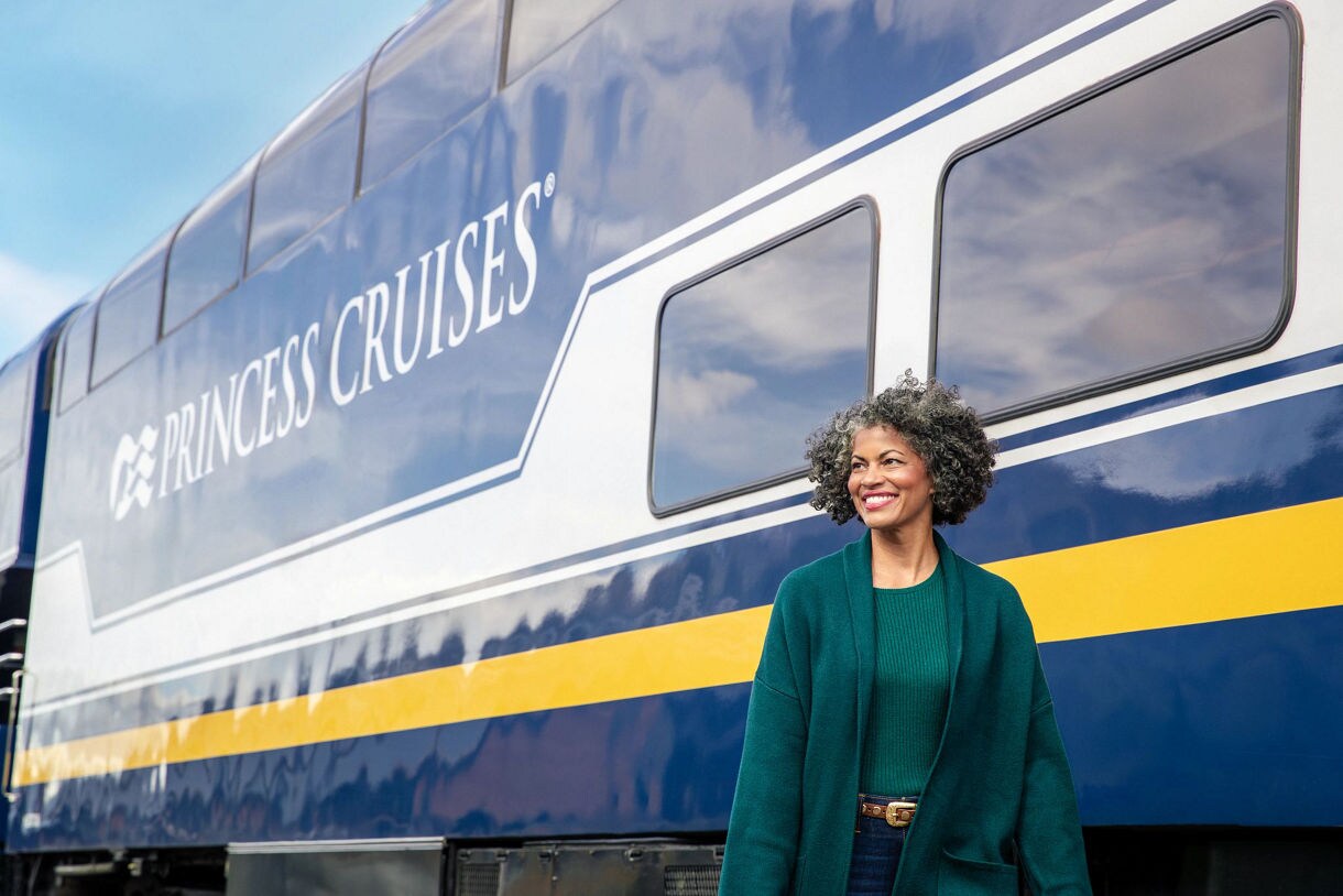 A woman with curly hair wearing a teal outfit stands smiling in front of a blue and yellow Princess Cruises train car