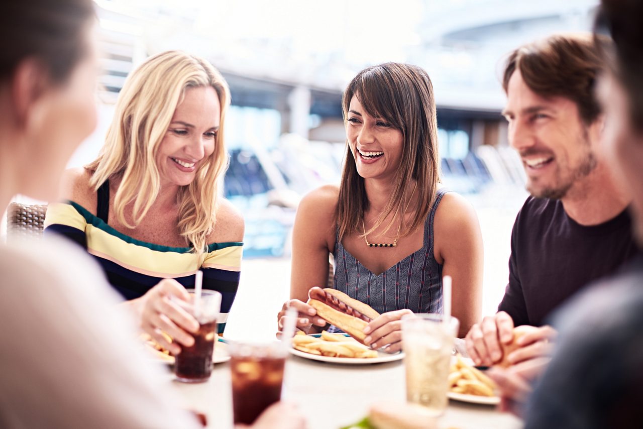 A group of friends laughing and eating burgers and fries together at an outdoor deck dining table.