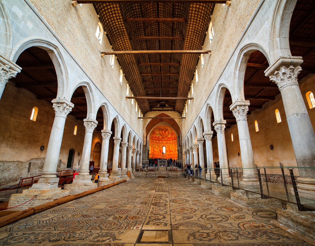Interior of an old basilica with tall stone columns, arched walkways, a wooden beamed ceiling and an expansive mosaic floor leading to a glowing orange frescoed apse.