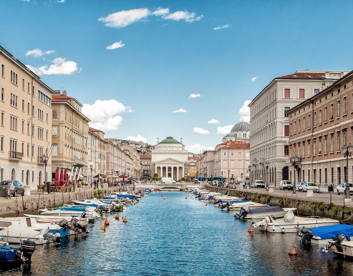 View of Trieste’s Canal Grande lined with pastel buildings and small boats, leading toward a white neoclassical church with a green dome under a bright blue sky with scattered clouds.