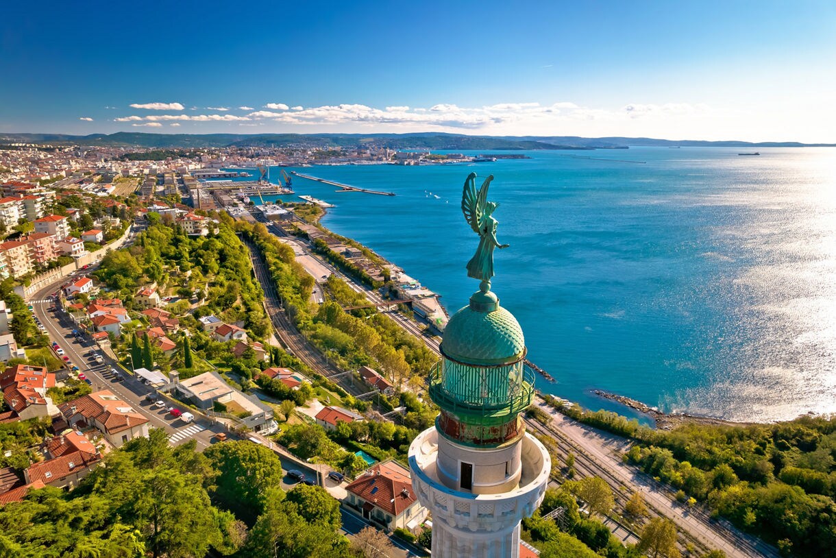 Aerial view of Trieste’s coastline with a tall lighthouse in the foreground topped by a green statue, overlooking blue Adriatic waters, rail lines and a spread of city buildings.