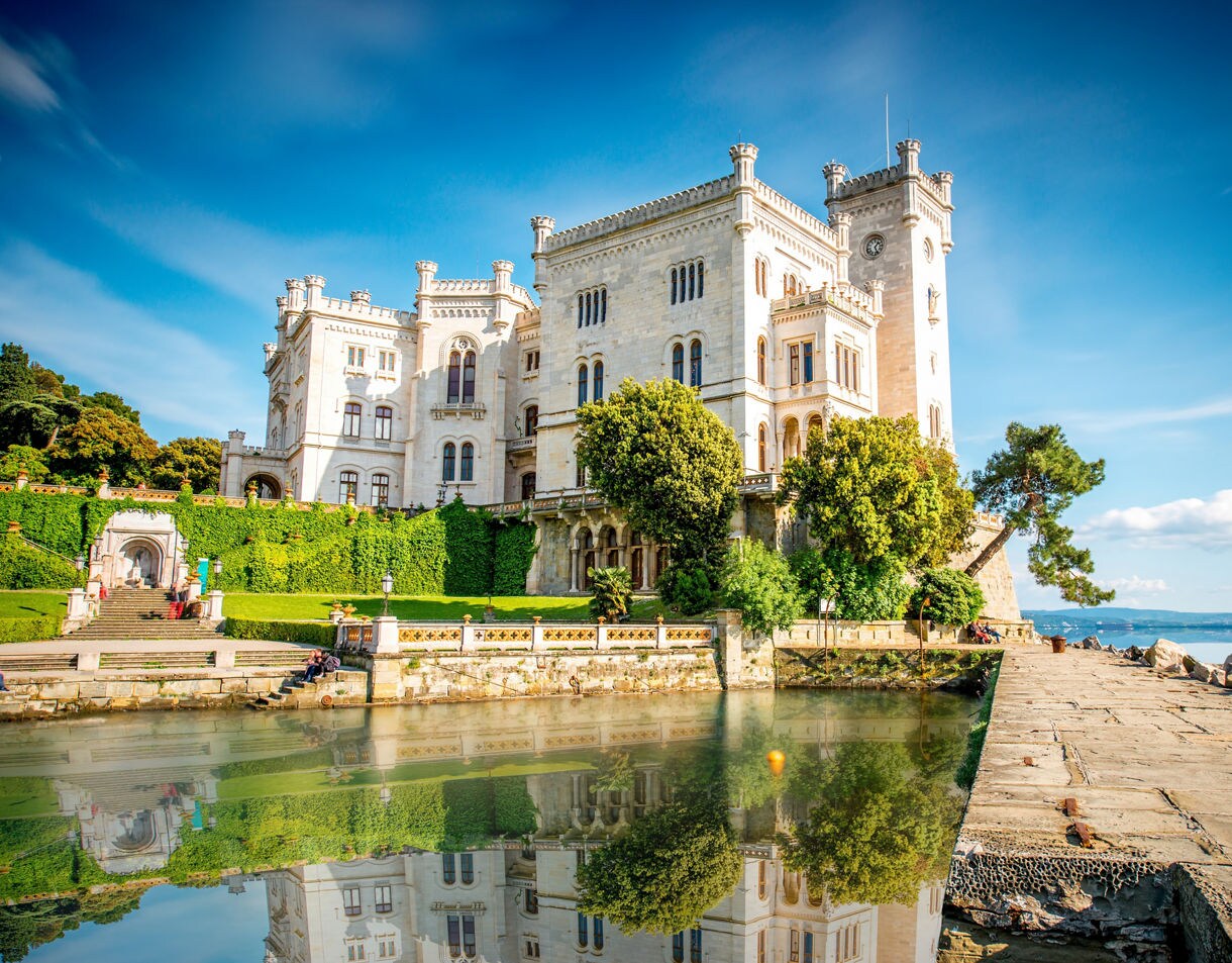 A grand white stone castle in Trieste, Italy, surrounded by gardens and reflected in a calm waterfront pool, with blue sky and distant coastline in the background.