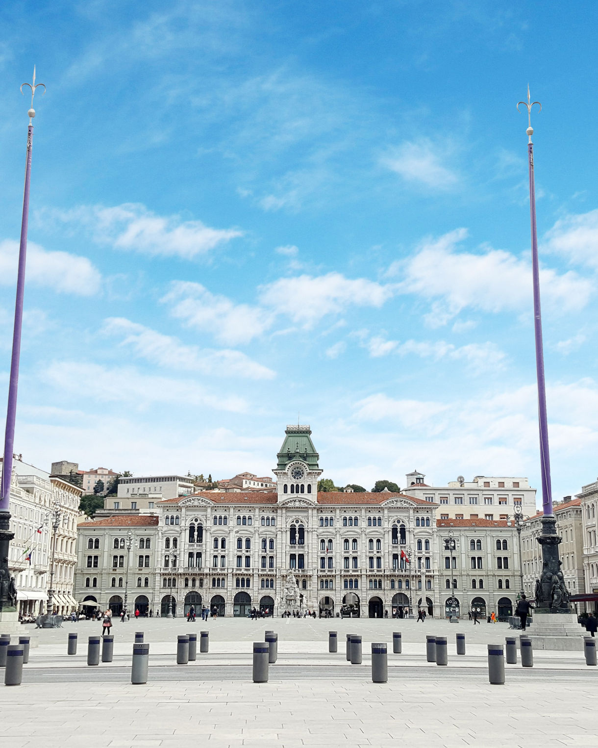 Wide view of Trieste’s Piazza Unità d’Italia featuring ornate white buildings, tall flagpoles, and a spacious stone plaza under a bright blue sky with scattered clouds.