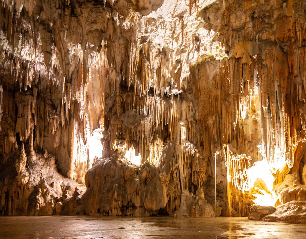 Interior of a large cave featuring illuminated stalactites and stalagmites in warm tones of brown and gold, with textured rock formations filling the frame.