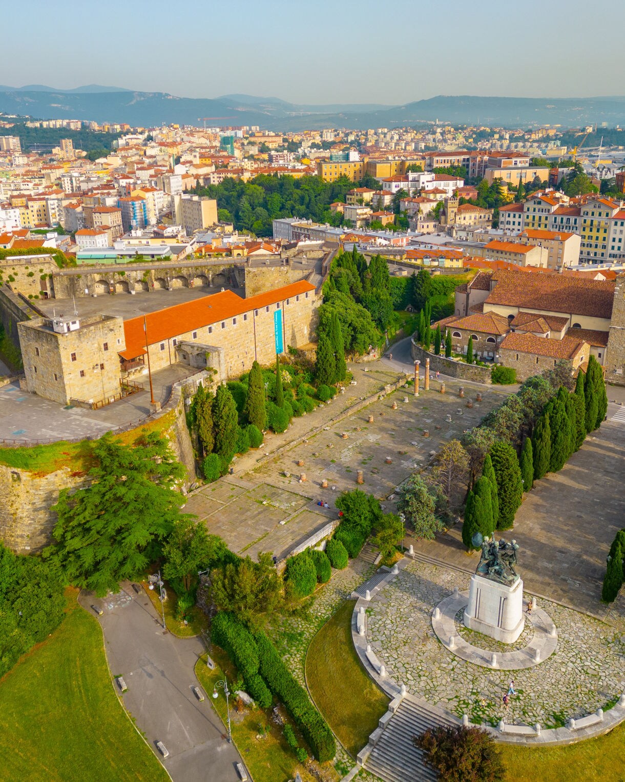 Aerial view of San Giusto Hill in Trieste featuring the stone fortress, the Basilica of San Giusto and tree-lined terraces, with the modern city and distant hills in the background.