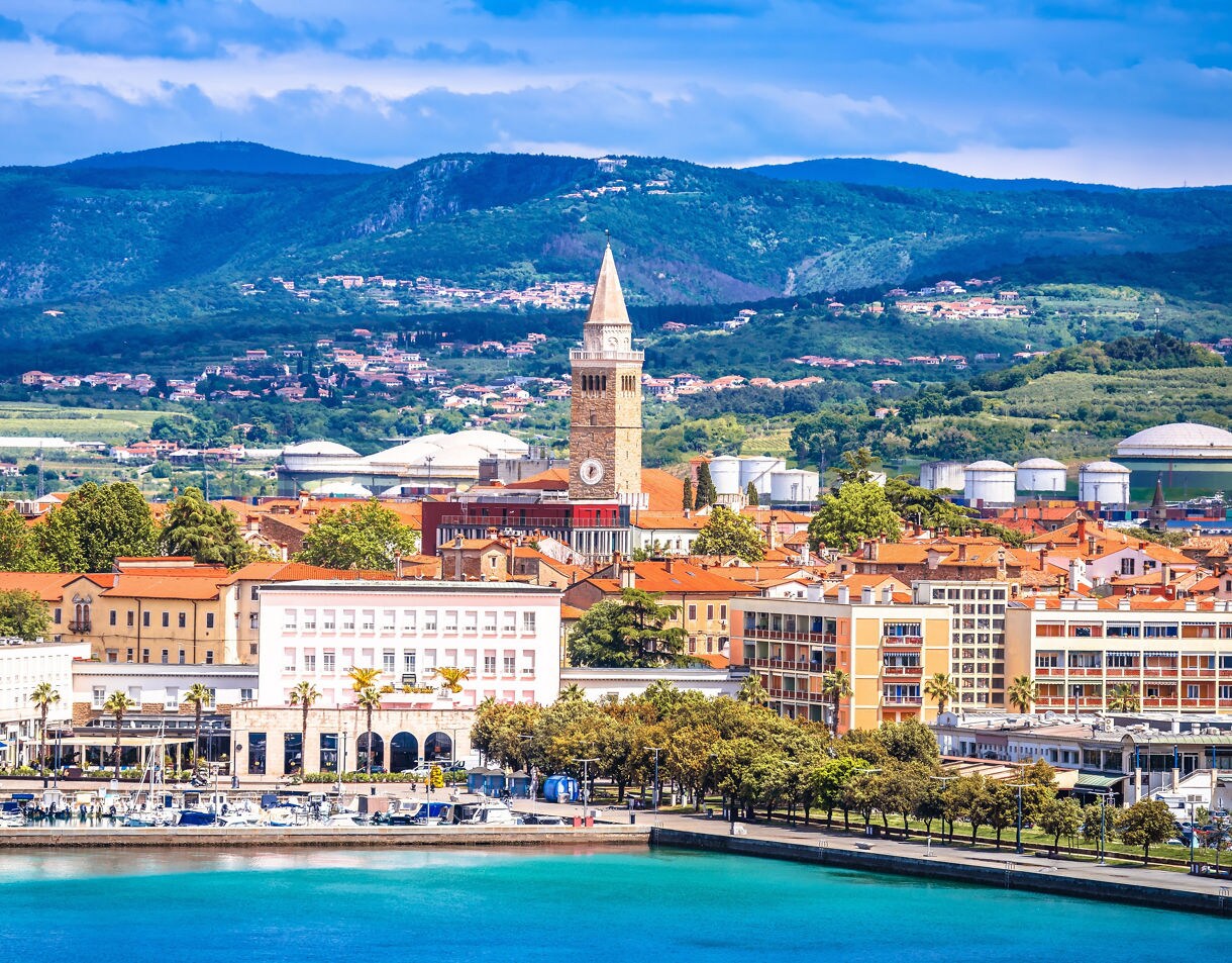 Aerial shot of a seaside city on the Adriatic featuring colorful buildings, a prominent bell tower and bright blue water backed by green hills.
