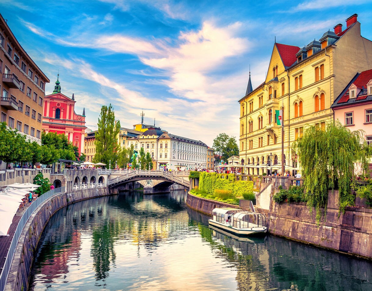 A vibrant view of Ljubljana’s riverfront with colorful historic buildings, a stone bridge, waterfront cafés and a tour boat reflected in the calm river under a bright sky.